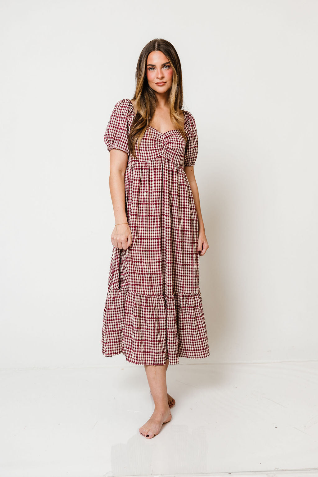 Model in the Maple Field babydoll midi dress in a rosy burgundy gingham check with short puff sleeves and a tiered skirt