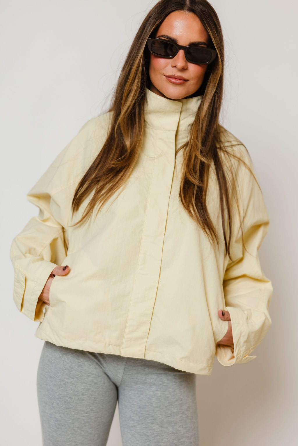 Model in the Drew oversized utility windbreaker jacket in pale buttercream yellow with a high zip collar, worn with grey pants