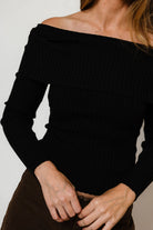Model in the Twyla ribbed knit off-the-shoulder sweater in black with long sleeves, worn with dark brown pants