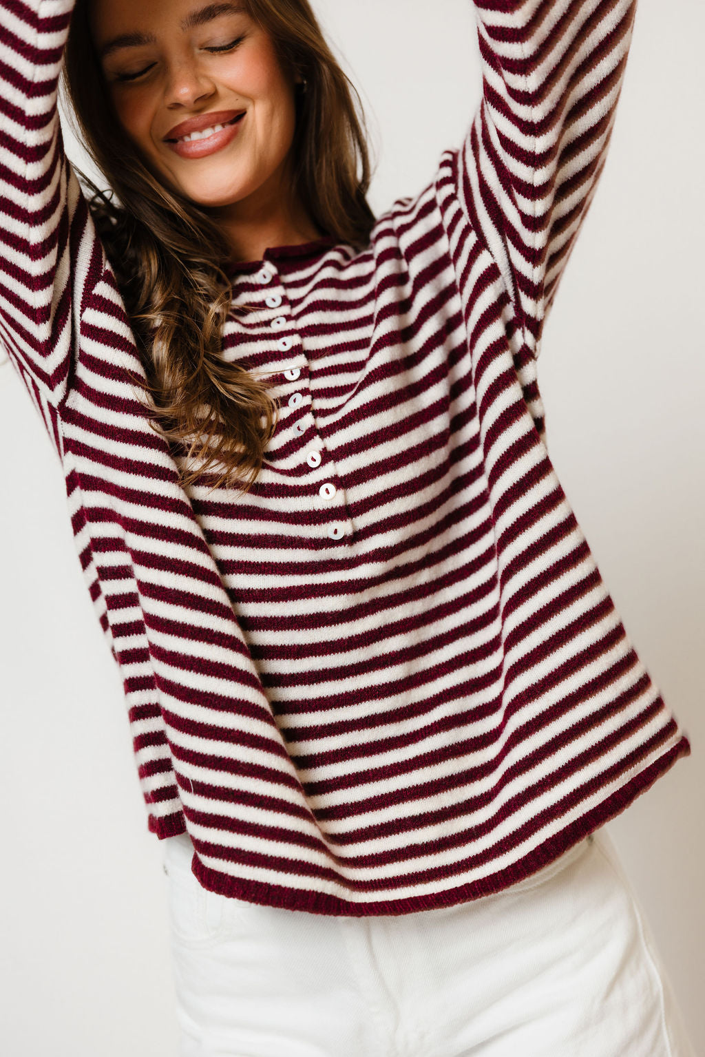 Model with arms raised in the Ever oversized henley sweater with wine and cream horizontal stripes, worn with white pants