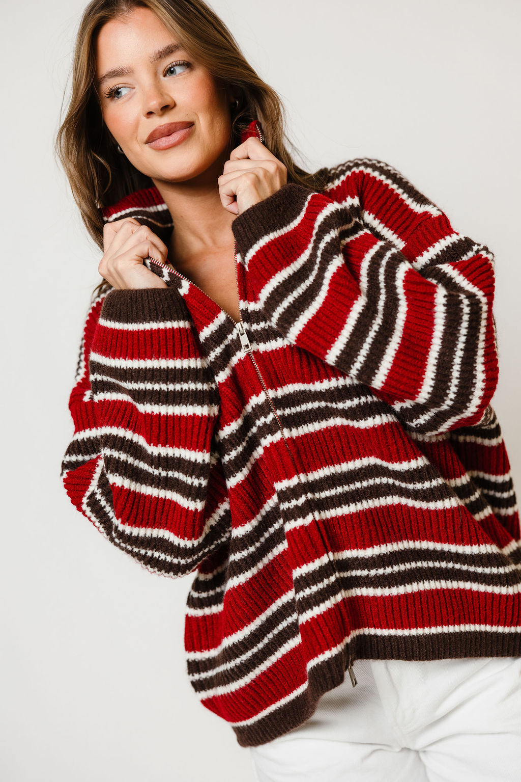 Model in the Kanna mock neck zip-up knit jacket with red, cream and dark brown stripes, worn with white pants