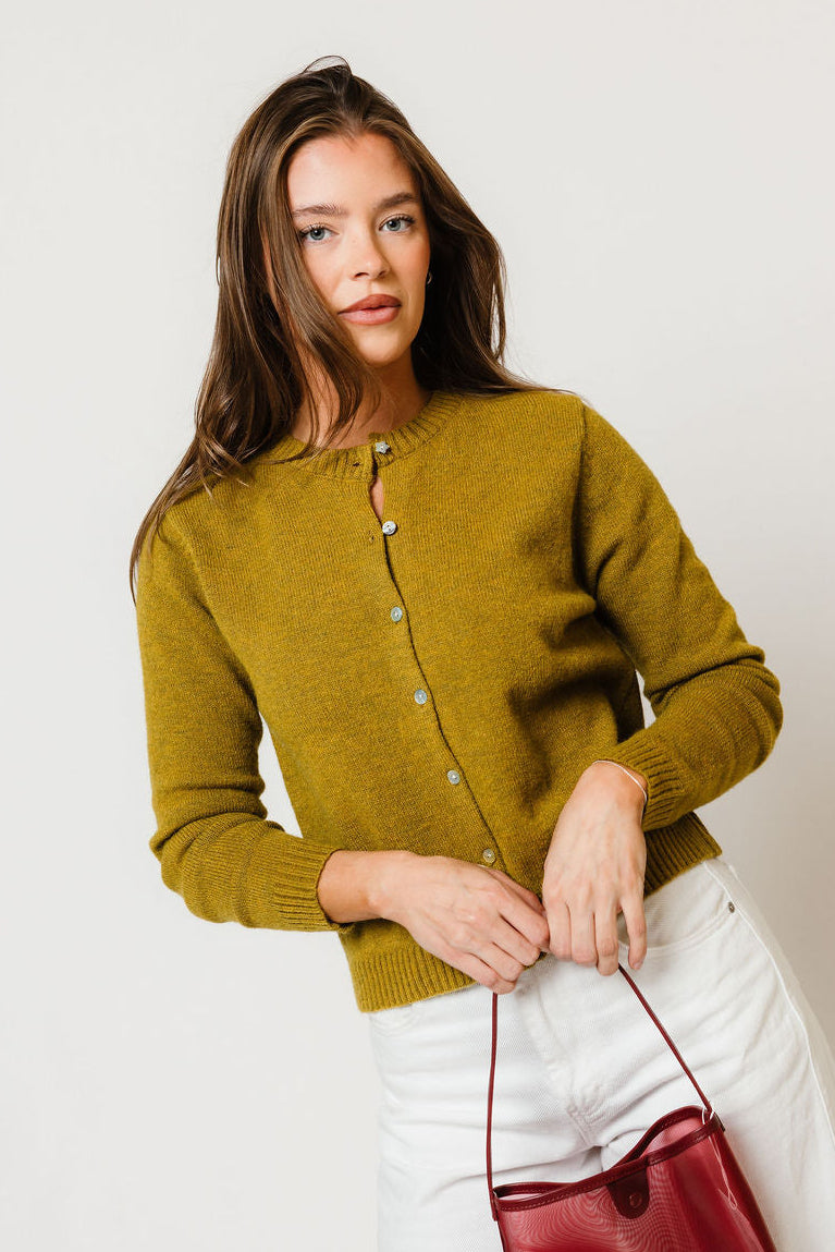 Model in the Elara button-front knit cardigan in moss olive green, worn with a white skirt and holding a red bag