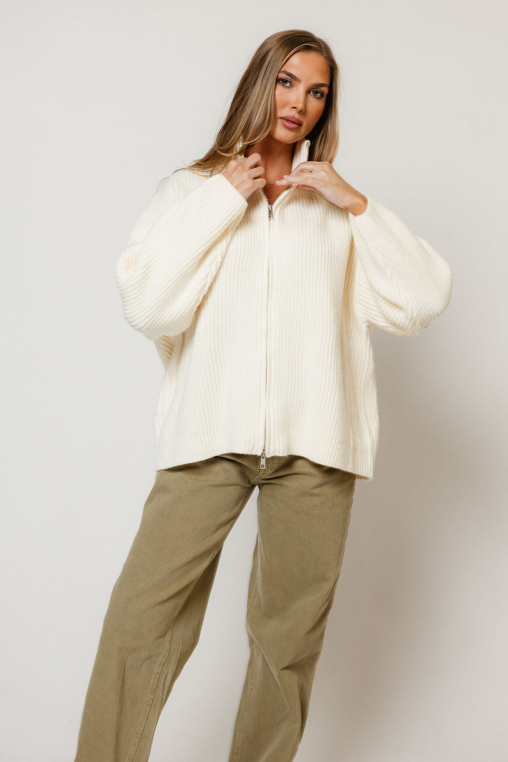 Model in the Kanna oversized mock neck zip-up knit jacket in cream, worn with olive pants